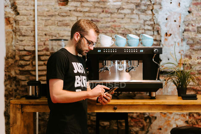 Vue latérale du barista masculin penché sur un comptoir en bois dans un café et un smartphone de navigation — Photo de stock
