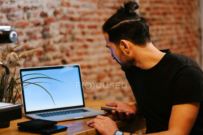 Vista lateral encantado barista masculino portátil de navegación mientras está sentado en el mostrador en la cafetería y el trabajo - foto de stock