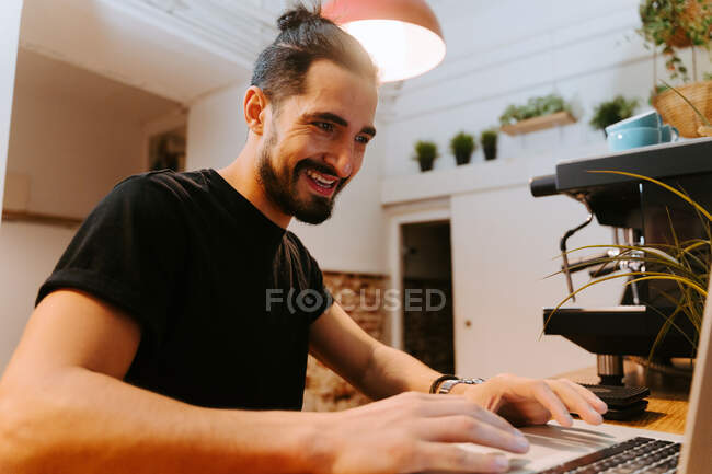 Low angle of delighted male barista browsing laptop while sitting at counter in coffee shop and working — Stock Photo