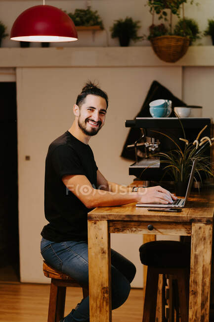 Vista lateral barista feliz navegando laptop enquanto sentado no balcão no café e trabalhando — Fotografia de Stock