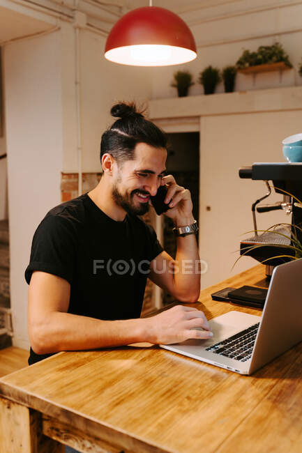 Sorria barista sentado no balcão de madeira com máquina de café e trabalhando enquanto fala no smartphone e usando laptop — Fotografia de Stock