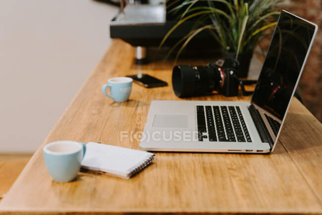 Lugar de trabajo del freelancer con netbook moderno y cámara fotográfica profesional colocada en mesa de madera con tazas de espresso y bloc de notas en la cafetería - foto de stock