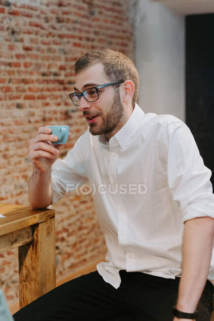 Male barista in white shirt and with small cup of espresso sitting at wooden counter in cafe and looking away — Stock Photo