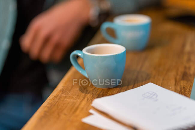 Pequeñas tazas para espresso colocadas en mostrador de madera con cuaderno en el fondo de la cosecha barista irreconocible trabajando en la cafetería - foto de stock
