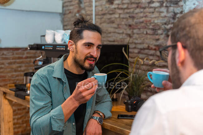 Delighted male baristas with small cups of espresso tasting prepared coffee while clinking mugs and looking at each other — Stock Photo
