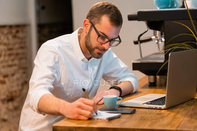 Мужчина-бариста сидит за деревянным столиком с ноутбуком и делает заметки в блокноте во время работы в кофейне — стоковое фото