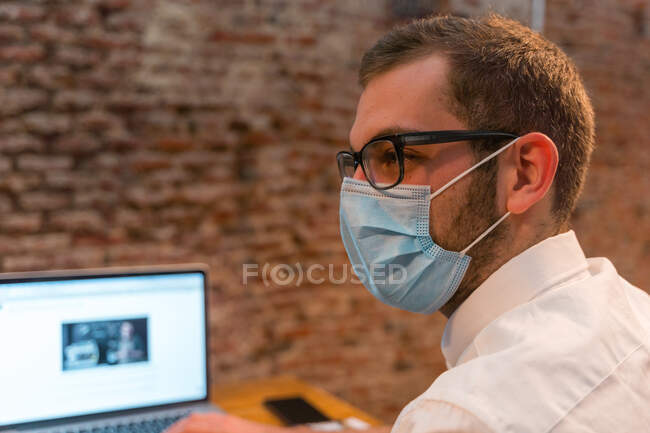 Side view of male barista in protective mask sitting at counter in coffee shop and working on laptop while looking away — Stock Photo