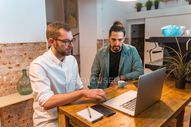 Varón baristas sentado en el mostrador con el ordenador portátil en la cafetería y hablando de problemas de trabajo - foto de stock