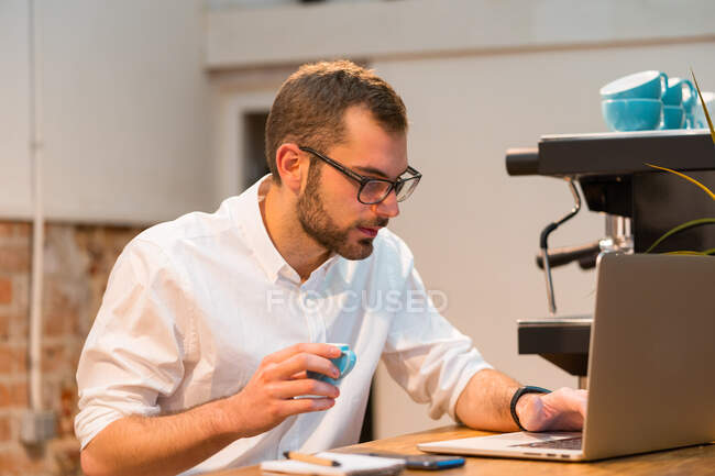 Орієнтований чоловік бариста сидить за дерев'яною стійкою з ноутбуком і робить нотатки в блокноті під час роботи в кав'ярні — стокове фото