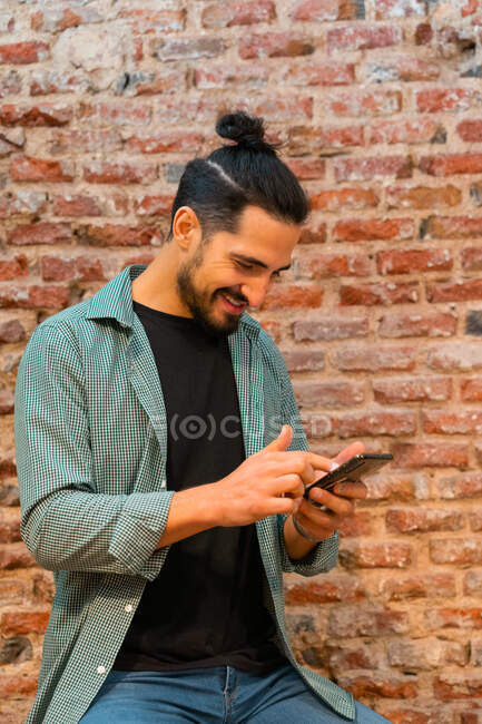 Side view of cheerful male barista browsing smartphone while working in loft styled cafe against shabby brick wall — Stock Photo