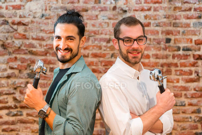 Vue latérale de baristas masculins joyeux avec des portafilters debout dos à dos et regardant la caméra sur le fond du mur de briques dans un café de style loft — Photo de stock