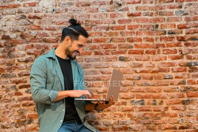 Vista lateral do barista masculino alegre que navega netbook enquanto trabalha no café estilo loft contra parede de tijolo gasto — Fotografia de Stock