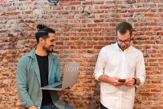 Zufriedene männliche Baristas arbeiten im Loft-Stil im Café zusammen, während sie Laptop und Smartphone benutzen — Stockfoto