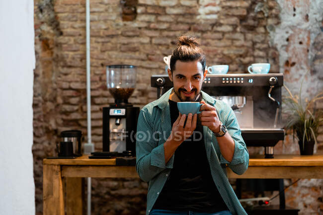 Barista mâle souriant assis dans un café et sentant boisson chaude aromatique tout en regardant la caméra — Photo de stock