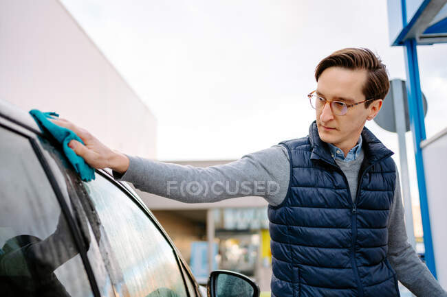 Сосредоточен молодой мужчина в повседневной одежде и очках, вытирая тряпкой транспортное средство, стоя на автомойке против облачного неба — стоковое фото