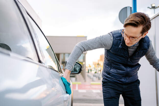 Focado jovem macho em roupas casuais e óculos limpando veículo com pano enquanto estava em pé na estação de lavagem de carro contra o céu nublado — Fotografia de Stock