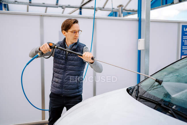 Hombre joven concentrado en traje casual y anteojos lavando parabrisas de coche con pistola de agua en la calle - foto de stock