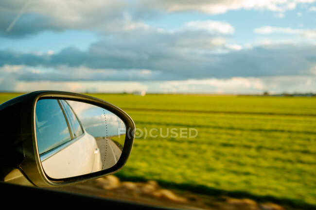 Through glass of side mirror of modern white car driving on asphalt road near endless green fields against cloudy blue sky in countryside — Stock Photo
