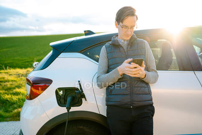 Концентрированный молодой водитель просматривает мобильный телефон, опираясь на современный автомобиль хэтчбэка, припаркованный возле зеленого поля во время заправки при солнечном свете — стоковое фото
