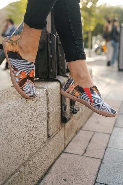 Cultivo piernas de mujer delgada con pantalones vaqueros y calzado de verano con mariposas relajantes en la calle de la ciudad - foto de stock