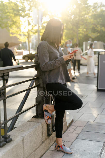 Vista lateral da fêmea irreconhecível em roupas casuais apoiadas em trilhos e mensagens no smartphone na cidade — Fotografia de Stock