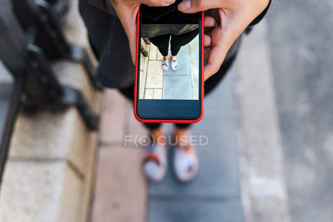 De cima de cultura blogueira fêmea anônima tirar fotos de pernas no smartphone enquanto em pé na rua na cidade — Fotografia de Stock