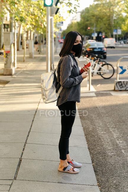 Vista laterale della femmina che indossa una maschera protettiva in piedi vicino al marciapiede mentre passeggia in città durante l'epidemia di coronavirus — Foto stock