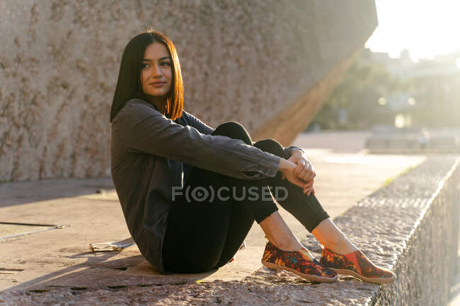 Side view of young peaceful female sitting on street and embracing knees while enjoying sunny day in city and looking away — Stock Photo