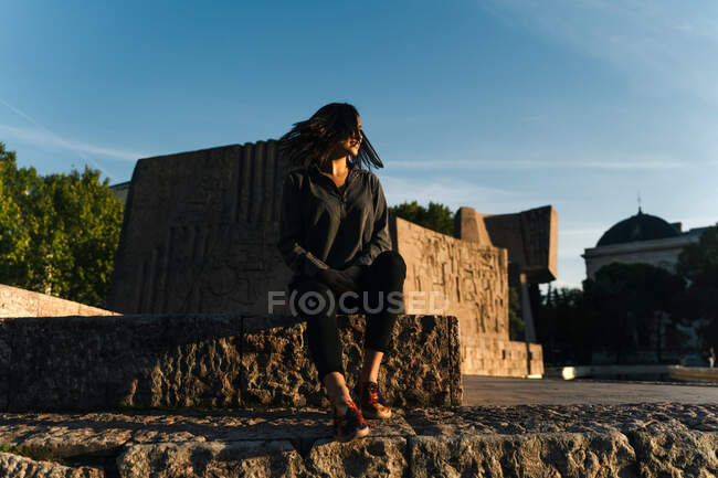 Низький кут безтурботної жінки з літаючим волоссям, що сидить біля кам'яного пам'ятника і дивиться далеко, насолоджуючись заходом сонця влітку — стокове фото