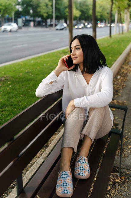 Mujer soñadora sentada en un banco de madera y hablando por teléfono móvil mientras se relaja y mira hacia otro lado - foto de stock