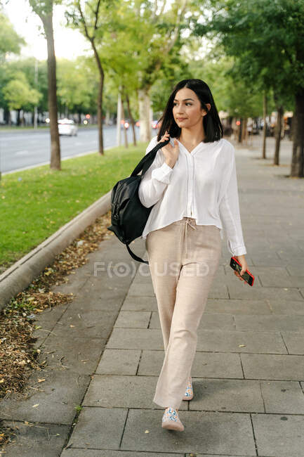 Молодая женщина в модном наряде и с сумочкой прогуливаясь по тротуару в городе и глядя в сторону, наслаждаясь прогулкой по выходным — стоковое фото