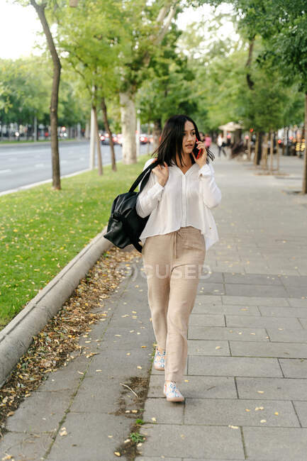 Joven mujer en traje de moda y con bolso caminando por el pavimento de la ciudad y mirando hacia otro lado mientras habla en el teléfono inteligente durante el paseo de fin de semana - foto de stock