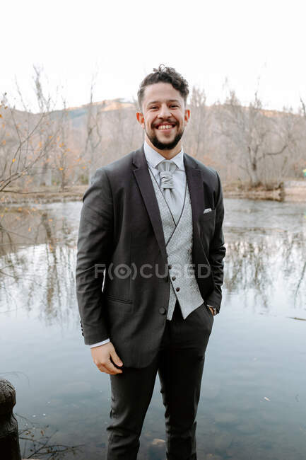 Cheerful young bearded groom in elegant suit smiling and looking at camera while standing on lake shore with hand in pocket on wedding day — Stock Photo