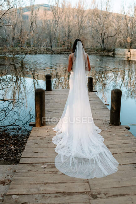 Back view of unrecognizable gorgeous young bride in elegant white wedding dress with long veil standing on wooden pier near lake in autumn forest — Stock Photo