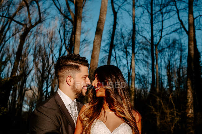Side view of young ethnic bride with long hair in white dress smiling and hugging groom with closed eyes while standing together near coniferous tree in forest — Stock Photo