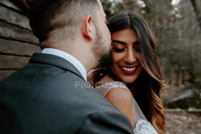 Side view of young groom in elegant suit kissing smiling ethnic bride while standing in nature on wedding day — Stock Photo