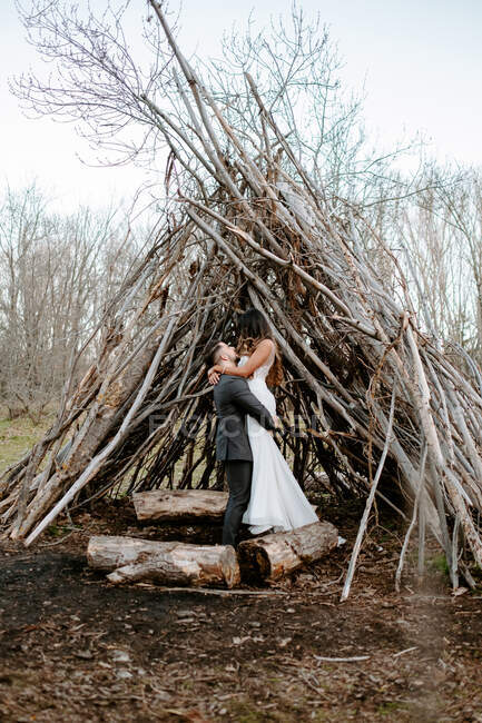 Vue latérale d'un jeune marié méconnaissable embrassant gracieuse mariée en robe de mariée blanche tout en se tenant dans la forêt près de brindilles hutte le jour nuageux — Photo de stock