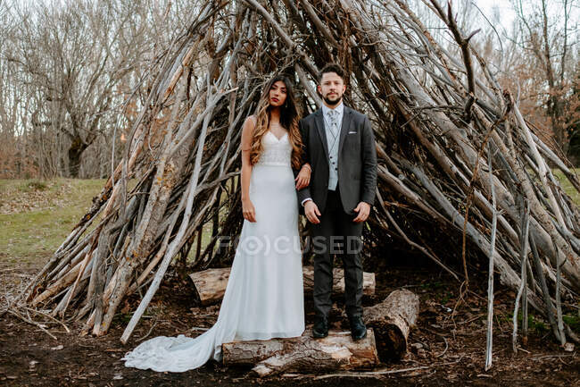 Young groom hugging graceful bride in white wedding dress while standing in forest near twigs hut on cloudy day — Stock Photo