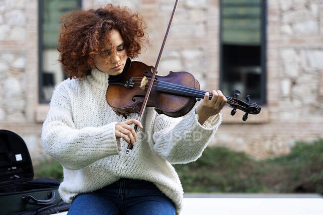 Young pretty female musician in casual wear sitting playing violin on boardwalk and looking at camera calmly in summer park — Stock Photo