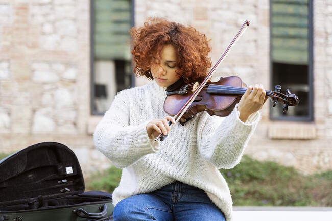 Молодая красивая женщина-музыкант в повседневной одежде играет на скрипке и спокойно смотрит в камеру, сидя на асфальтированной улице — стоковое фото