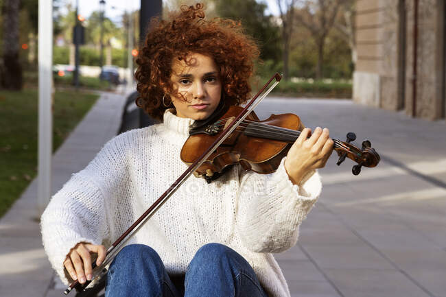 Junge hübsche Musikerin in Freizeitkleidung spielt Geige und blickt gelassen in die Kamera, während sie auf gepflasterter Straße sitzt — Stockfoto
