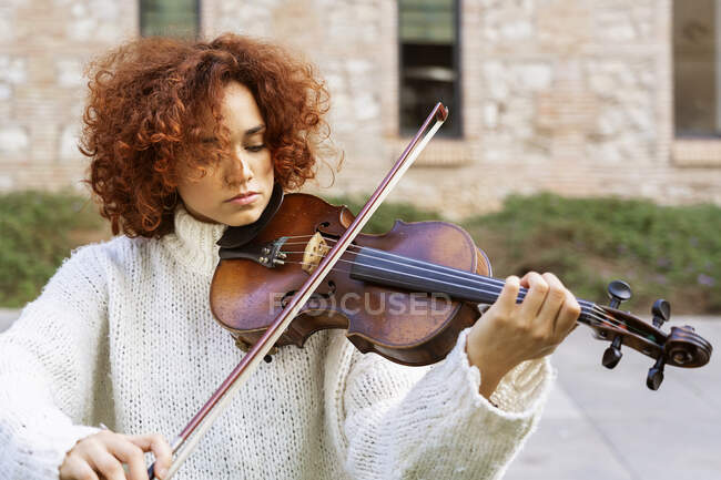 Молодая красивая женщина-музыкант в повседневной одежде играет на скрипке и спокойно смотрит в камеру, сидя на асфальтированной улице — стоковое фото