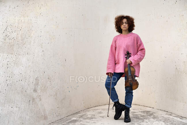 Рыжая музыкантка без эмоций, стоящая в повседневной одежде с закрытыми глазами и держащая акустическую скрипку на белой бетонной стене — стоковое фото