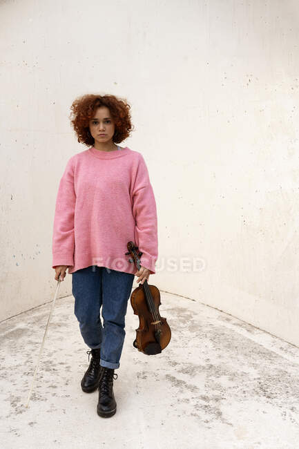 Musicista donna rossa senza emozioni a corpo intero in abito casual in piedi con gli occhi chiusi e tenendo il violino acustico contro il muro di cemento bianco — Foto stock