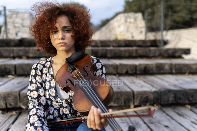 Young pretty female musician in casual wear sitting playing violin on boardwalk and looking at camera calmly in summer park — Stock Photo