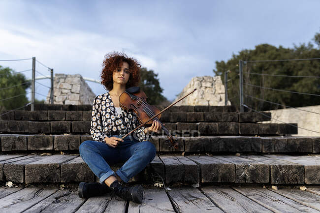 Ganzkörperjunge hübsche Musikerin in Freizeitkleidung sitzt mit Geige auf der Uferpromenade und blickt gelassen in die Kamera im Sommerpark — Stockfoto