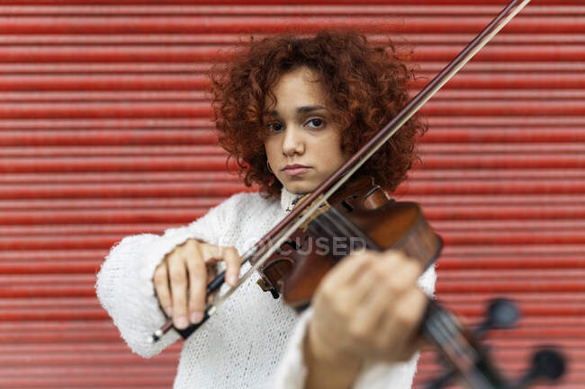 Glücklich schöne professionelle Musikerin in weißem Pullover, die akustische Geige spielt und mit zahmem Lächeln vor roter Wand in die Kamera blickt — Stockfoto