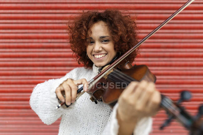 Feliz bonito músico feminino profissional em camisola branca tocando violino acústico e olhando para a câmera com sorriso de dente contra a parede vermelha — Fotografia de Stock