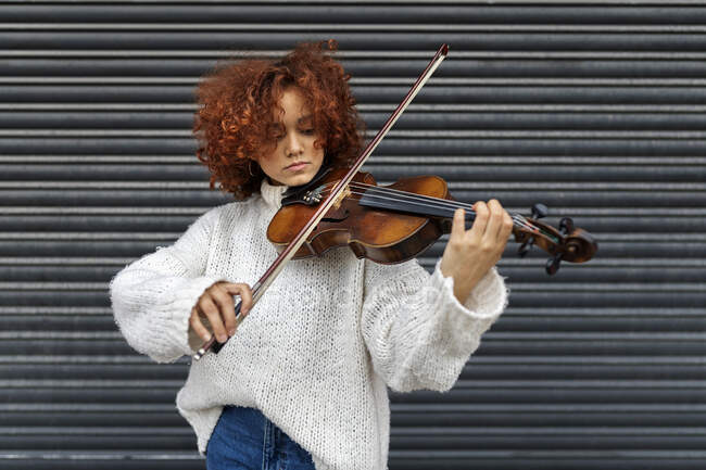 Wistful young professional female musician with red curly hair wearing casual sweater standing with acoustic violin against building wall and looking at camera — Stock Photo