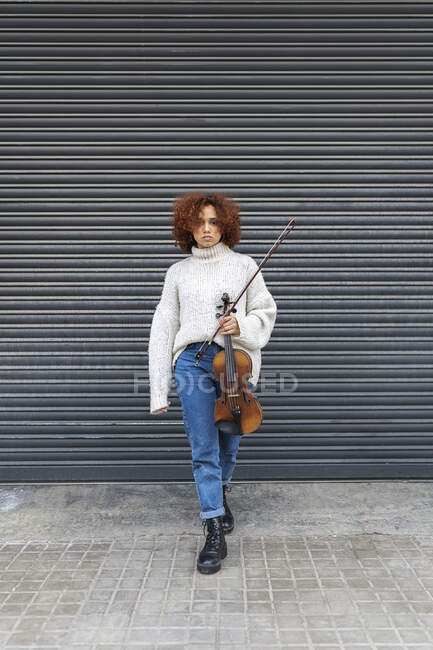Músico profesional joven y melancólico con el pelo rizado rojo usando suéter casual de pie con violín acústico contra la pared de la construcción y mirando a la cámara - foto de stock